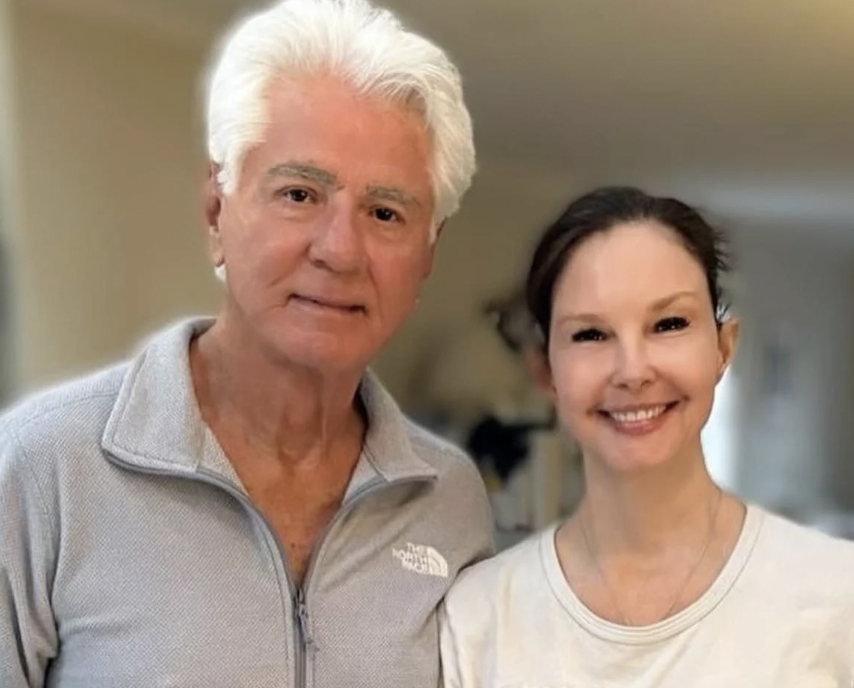 A man with white hair and a light gray North Face jacket stands next to a woman with brown hair pulled back, wearing a white t-shirt. Both are smiling at the camera in a warmly lit indoor setting.